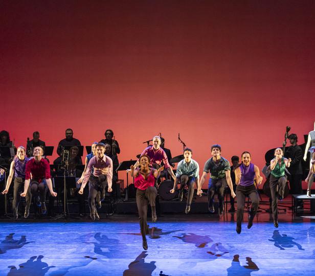 A vibrant stage performance featuring 舞者 energetically leaping into the air. 在后台, a band with various musicians is visible, contributing to the dynamic atmosphere of the show. The background is a bold red, enhancing the excitement and energy conveyed by the performers. The floor of the stage is adorned with blue patterns, suggesting 运动 and flow, which complements the dynamic poses of the 舞者.