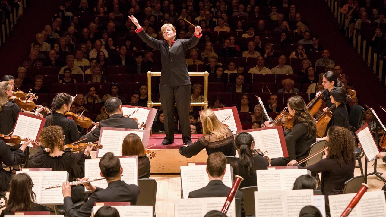 茱莉亚管弦乐团正在演奏, 由马林·阿尔索普带队. The conductor, positioned in the center, 穿着深色西装，高举双臂, baton in h和, expressing passion 和 comm和. The musicians, 专注地演奏乐器, 在音乐厅的舞台上以传统管弦乐的方式排列. 观众 fills the theater, 在灯光明亮、盛大的场地见证这一盛事, 这会增加正式和有声望的气氛吗