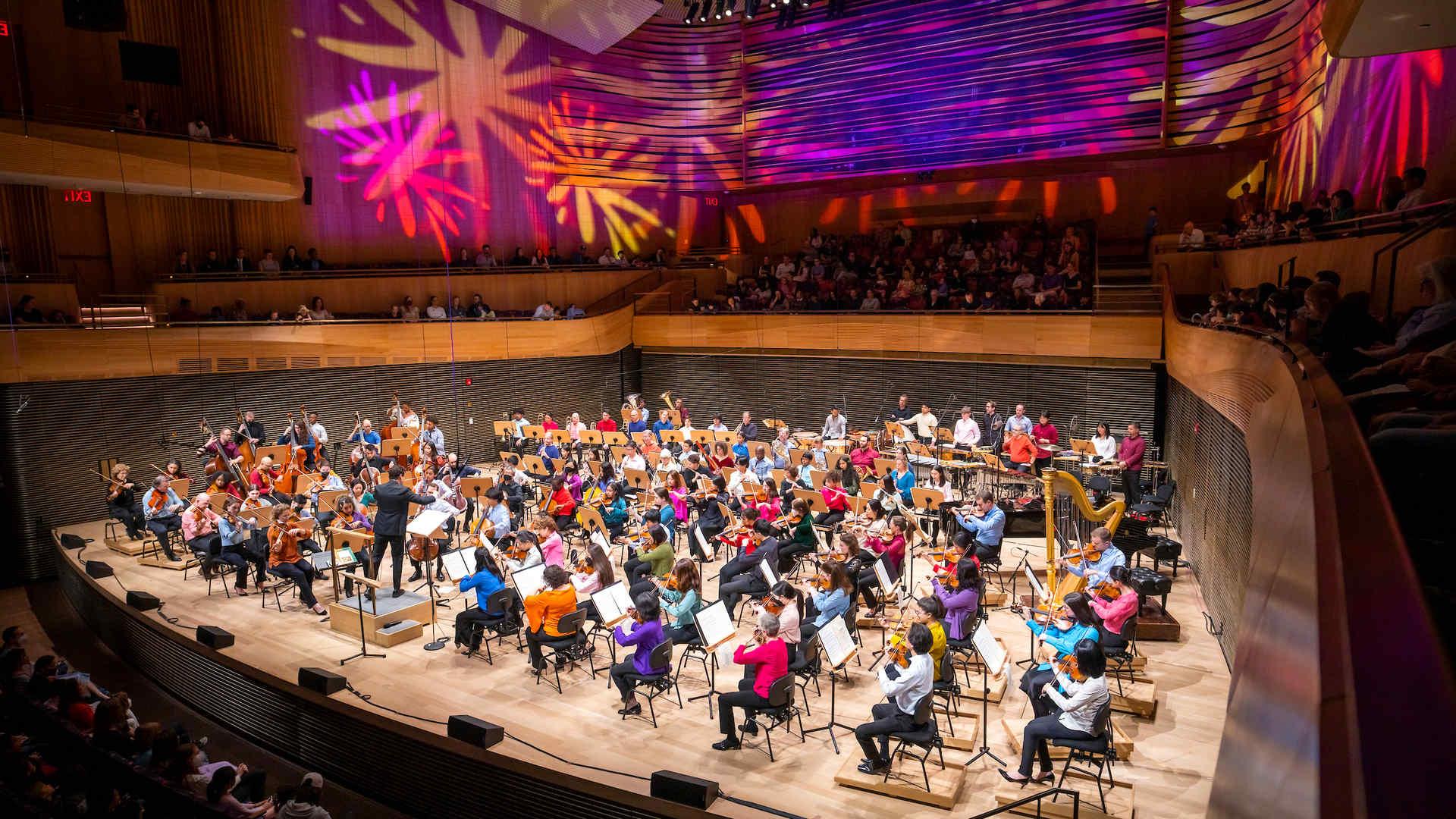 The photo captures a vibrant orchestral performance in progress inside a modern concert hall. 音乐家 are seated in a semi-circular arrangement on stage, surrounded by wood-paneled walls that reflect the colorful, abstract light patterns projected above them, creating a dynamic 和 festive atmosphere. A conductor is visible at the forefront, leading the orchestra. 观众, seated in the curved balconies 和 the main floor, is attentively watching the performance in a well-lit, 拥挤的场所.