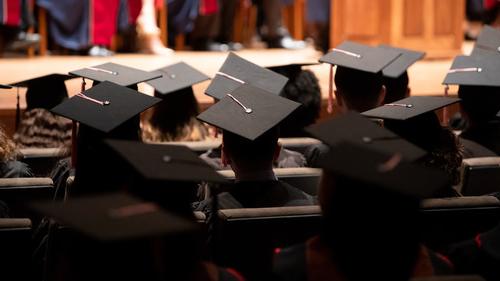 A group of graduates during a commencement ceremony. They are wearing traditional black caps and gowns. 设置 is Alice Tully Hall with rows of seating filled with the graduates. The angle of the photo suggests it’s taken from the back of the hall, focusing on the back of the graduates' heads and their mortarboards, with one in the foreground serving as a focal point.