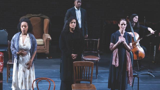 A group of actors and a cellist perform on stage, evoking deep emotions with their expressions. Three actors stand in the foreground, each expressing intense feelings through their posture and facial expressions, while another actor and a cellist are positioned in the background. The set includes various chairs and an armchair, creating a minimalist yet poignant scene.